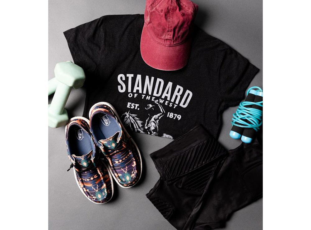 A heather gray t-shirt,  conceal carry leggings in black, and women’s hazer print casuals in multicolor prism posed with workout equipment. 